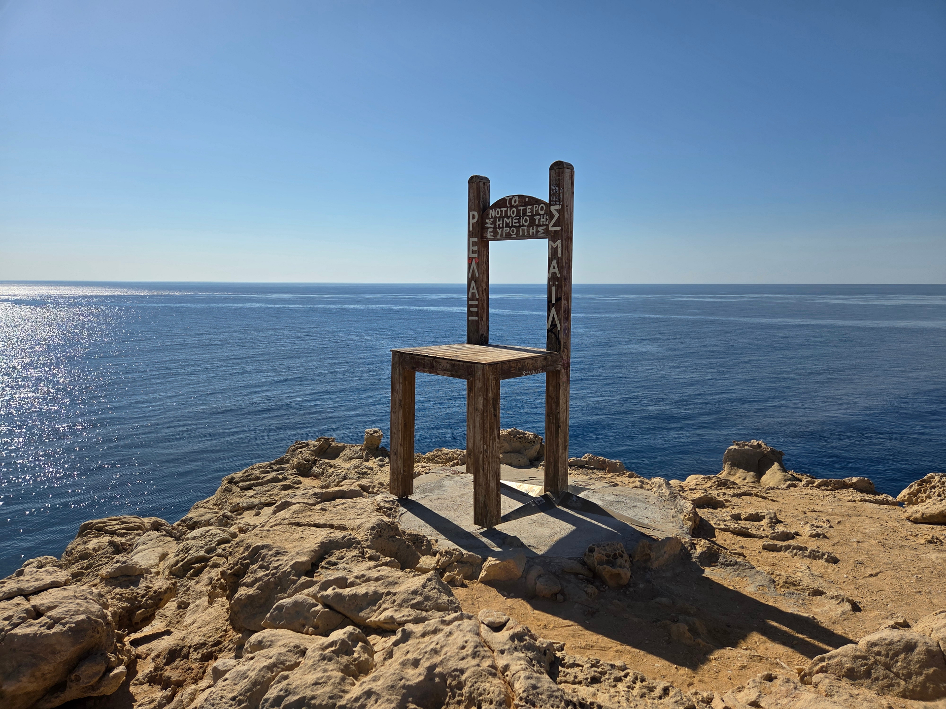 Silla de madera gigante mirando al mar infinito desde un acantilado, bajo un cielo azul despejado